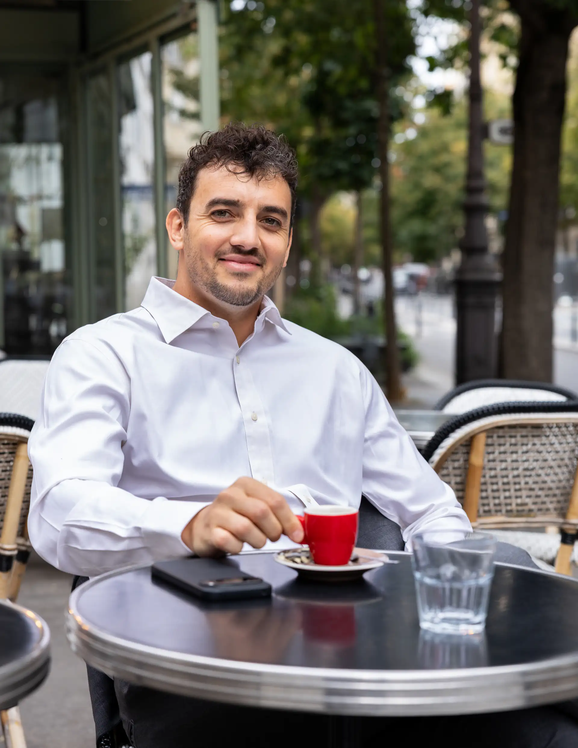 Théo Mével assis en chemise blanche à une table en terrasse, une main près d’une tasse rouge.