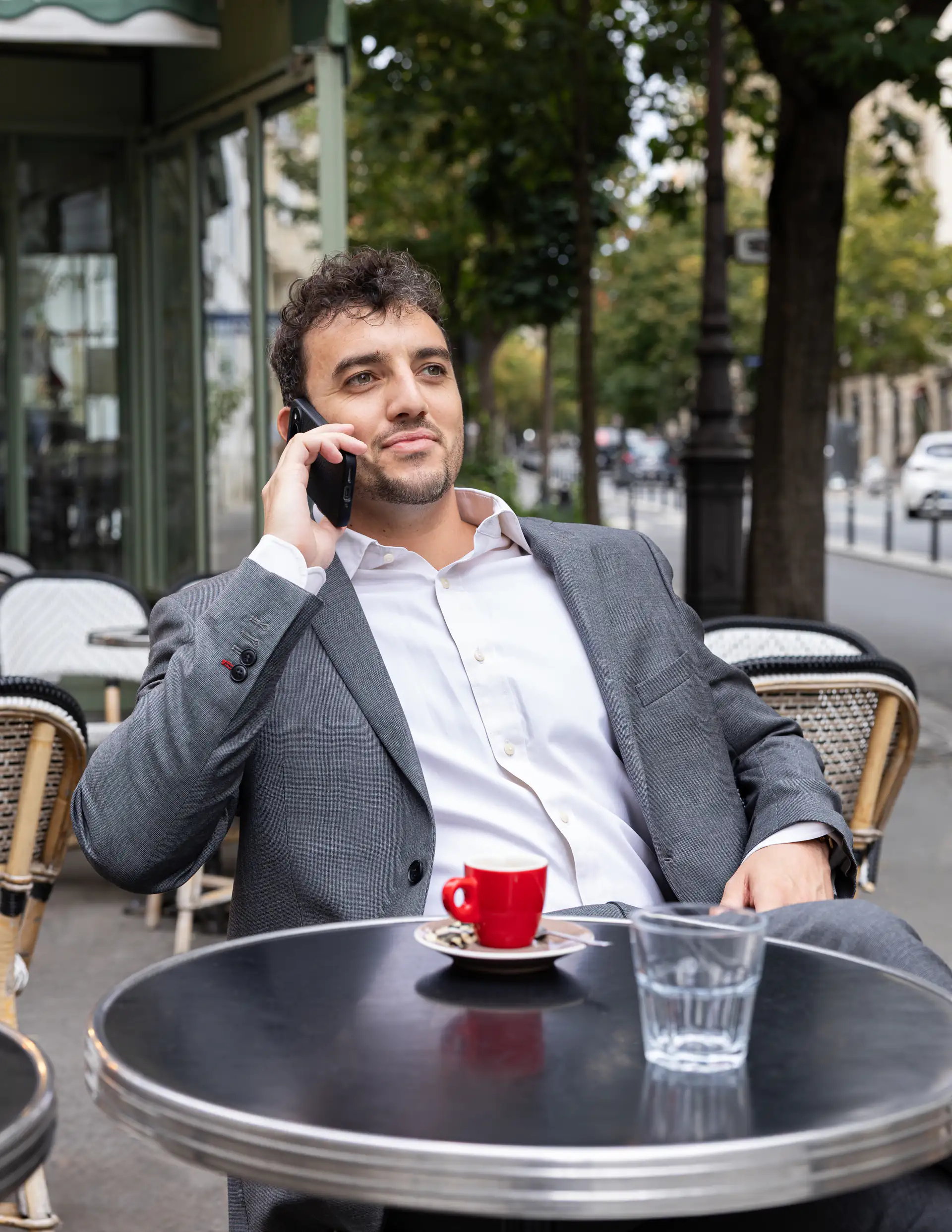 Théo Mével assis à une table en terrasse, téléphone à l’oreille, avec une tasse rouge devant lui.